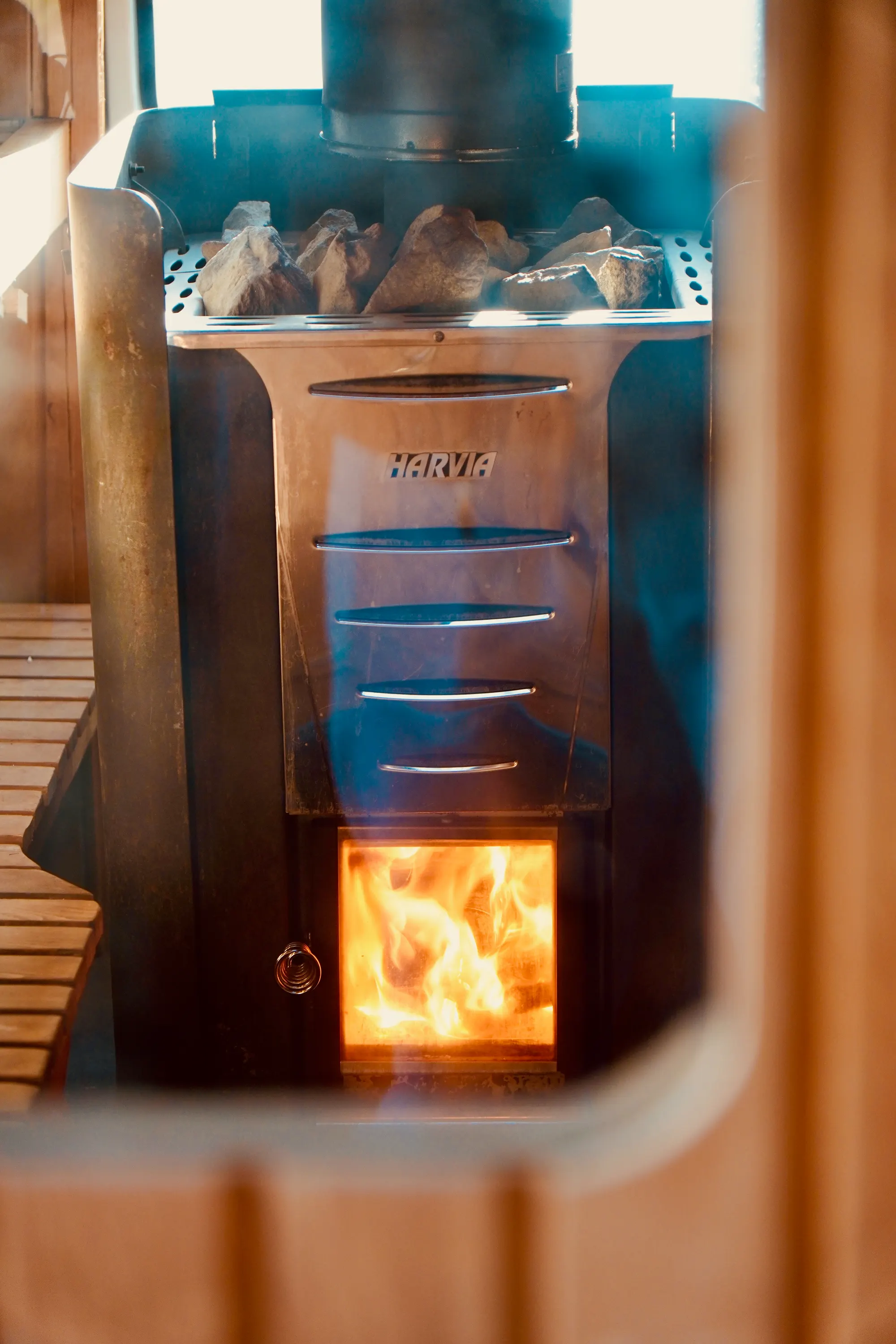 Nahaufnahme des Ofens mit Steinen obenauf und Flammen im Feuerraum, durch eine Öffnung in der Holzwand gesehen.
