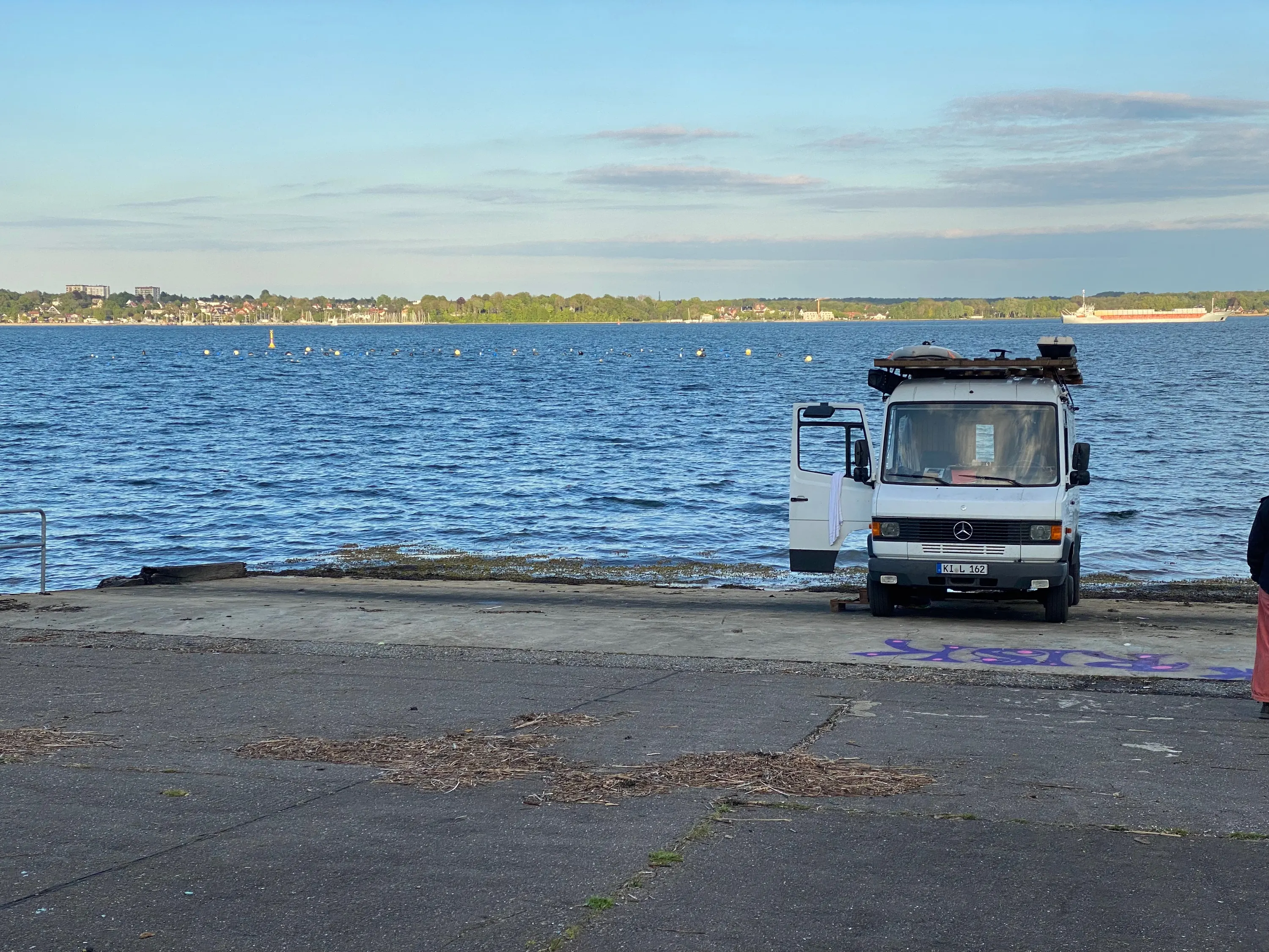 Der Saunabus mit offener Tür an der Wasserkante, dahinter die Bucht und das ferne Ufer.