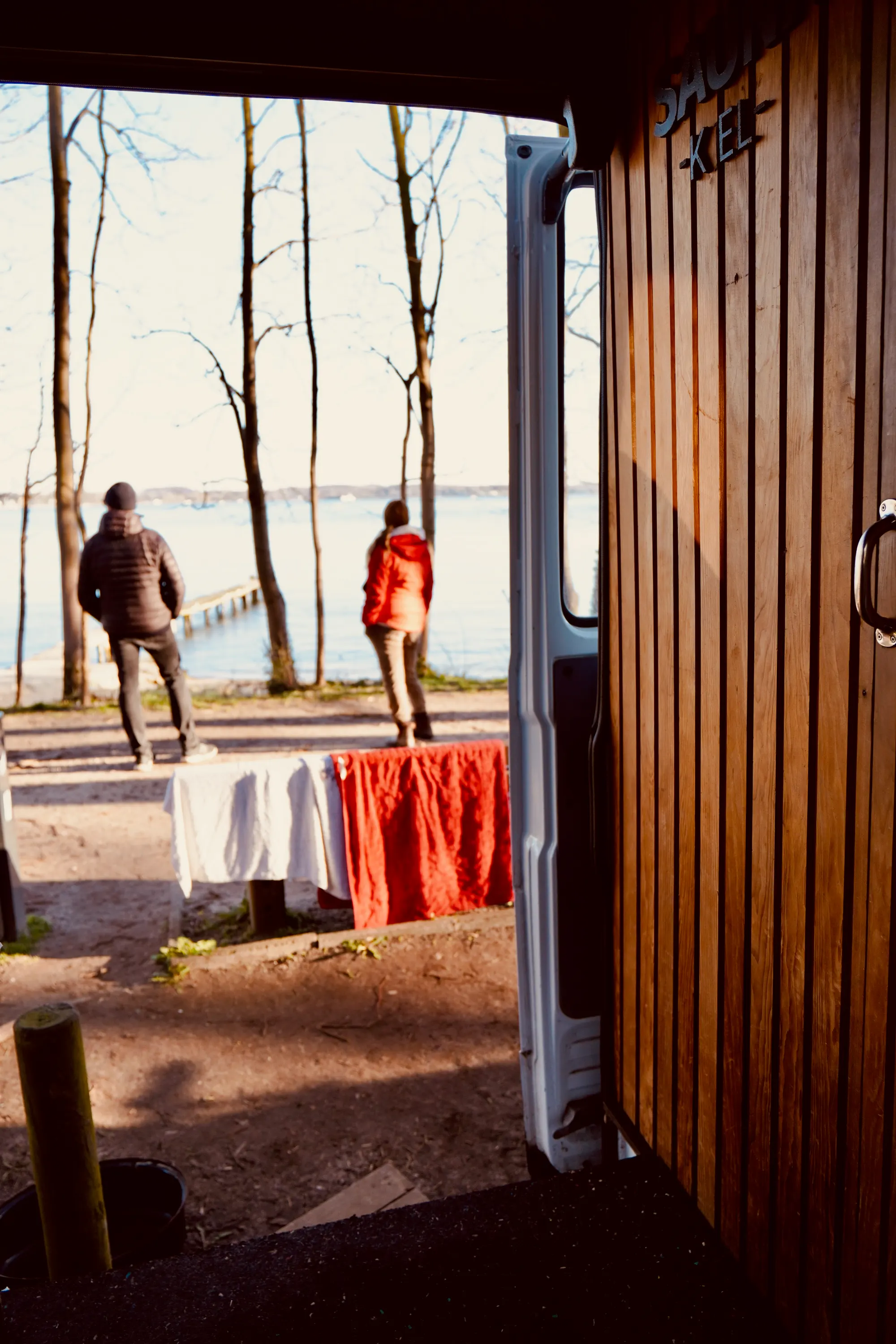 Blick aus der offenen Bustür auf kahle Bäume und das Wasser; zwei Personen von hinten am Ufer, Handtücher hängen zum Trocknen.