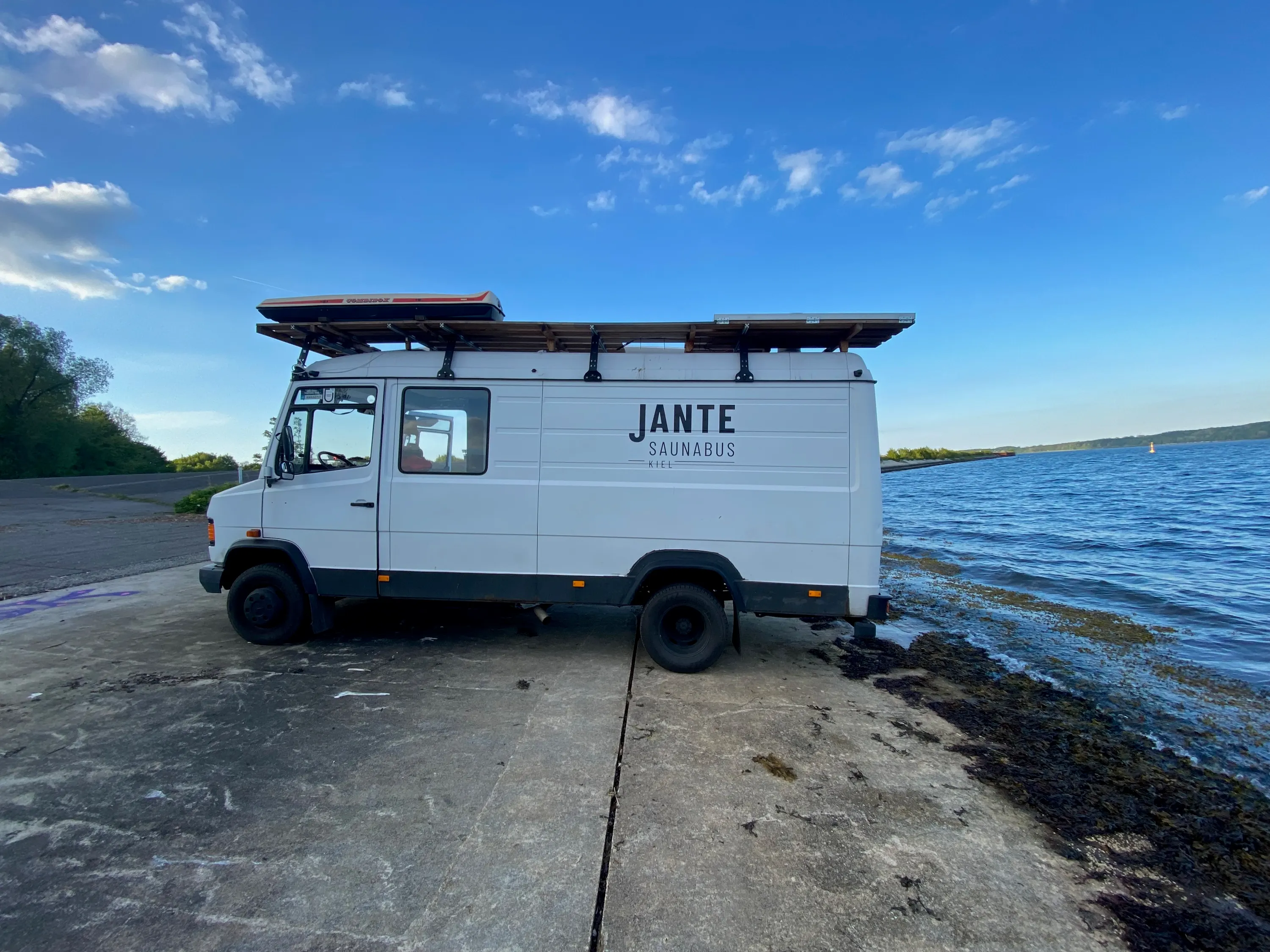 Der Saunabus steht seitlich auf einer betonierten Slipanlage direkt am Wasser unter blauem Himmel.