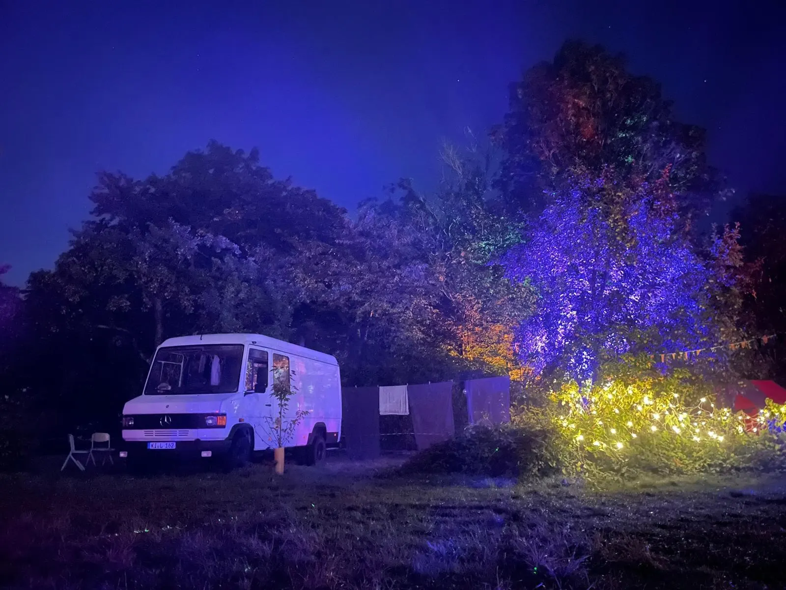 Der Saunabus bei Nacht auf einer Wiese, Bäume in blauem und violettem Licht, eine gelbe Lichterkette leuchtet.